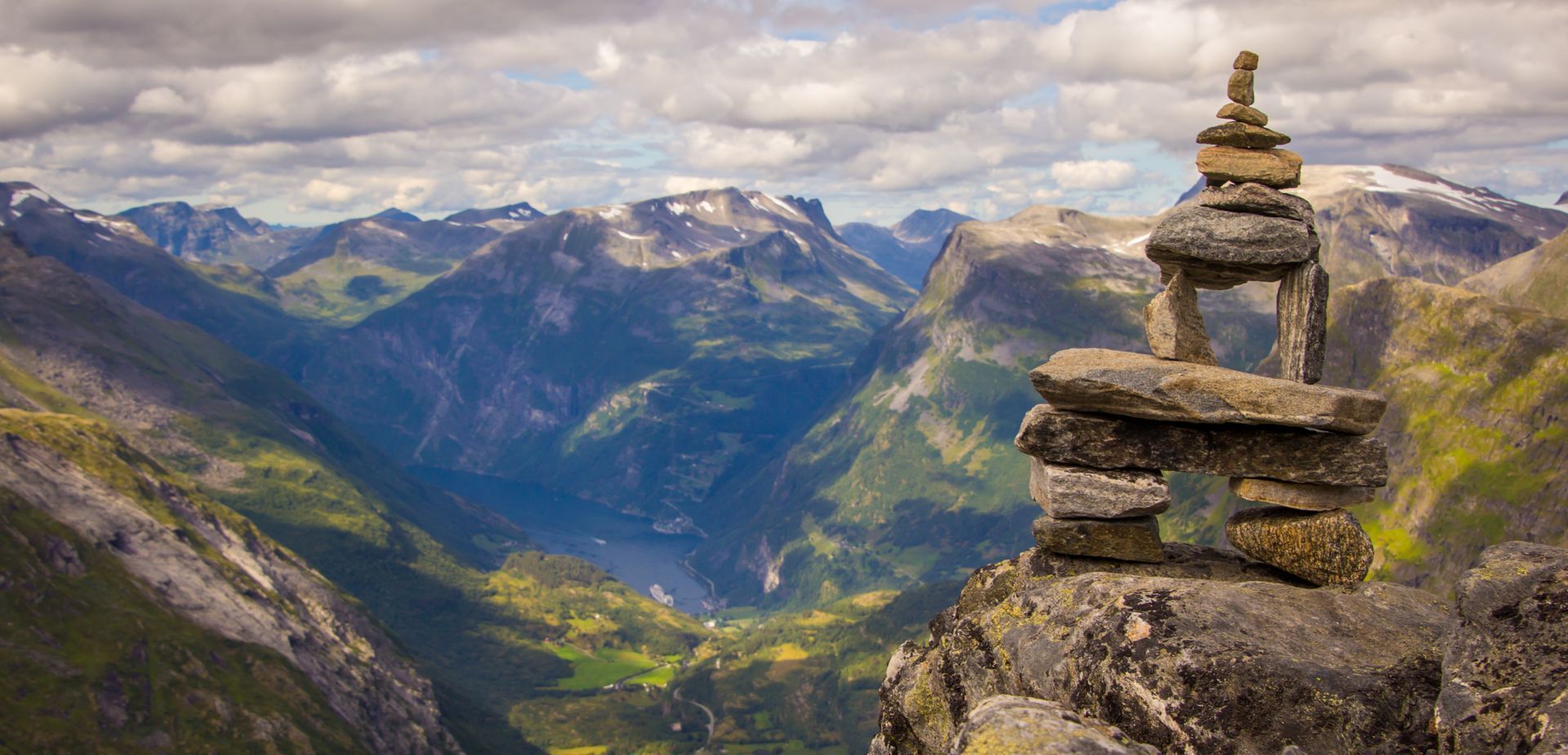 Stone cairn on a mountaintop overlooking a valley with mountains under a cloudy sky.
