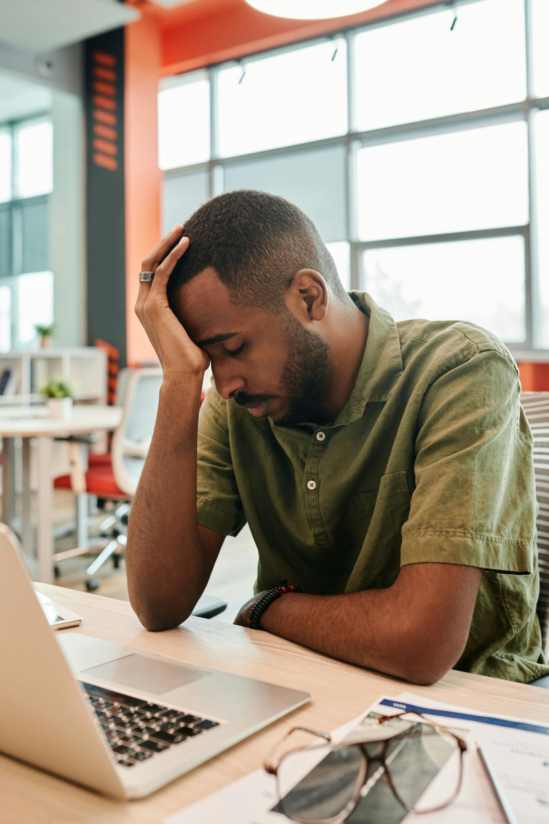 Man with hand on forehead, looking down at a laptop in an office setting. Appears stressed.