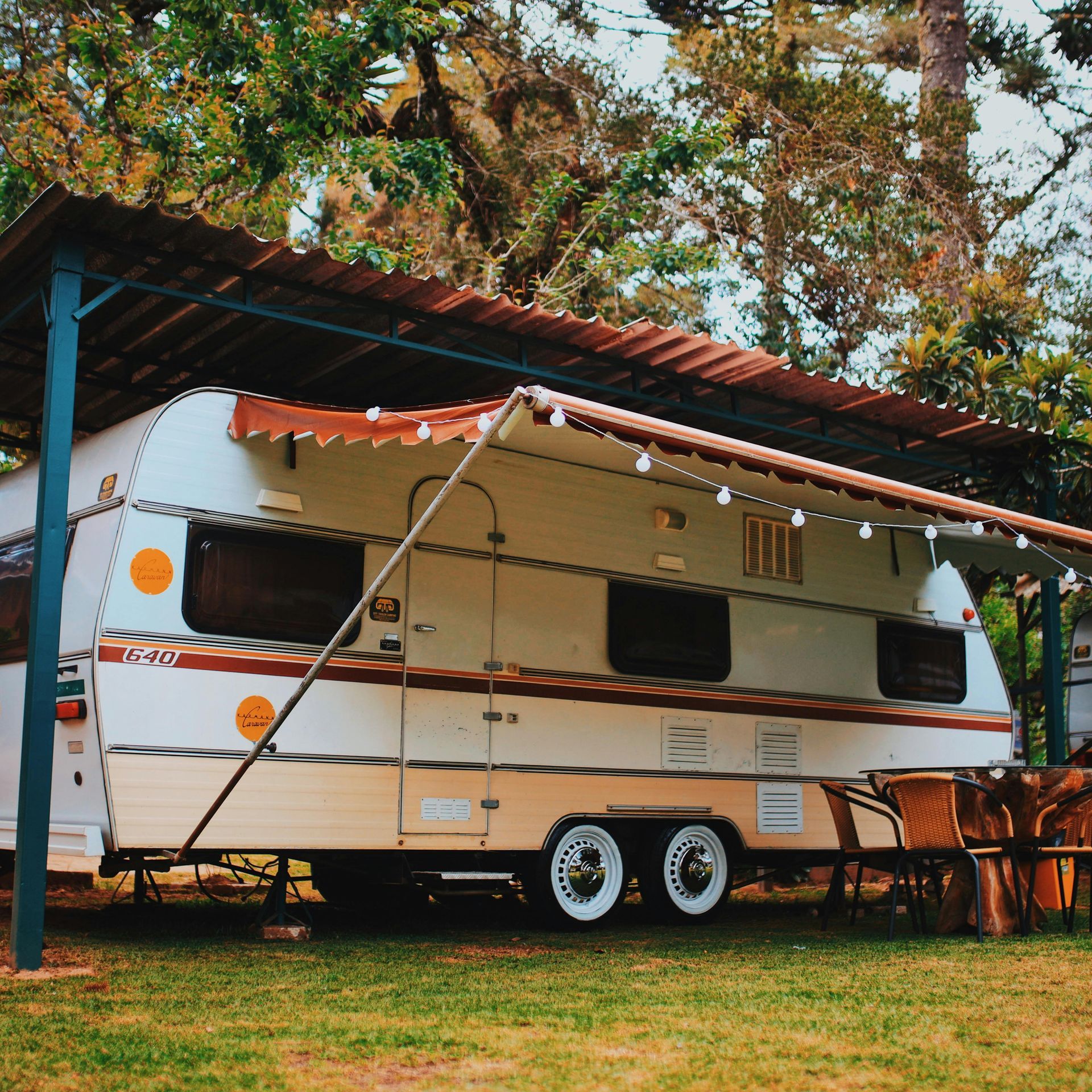 Camper trailer with awning, under a corrugated metal roof, parked on grass.