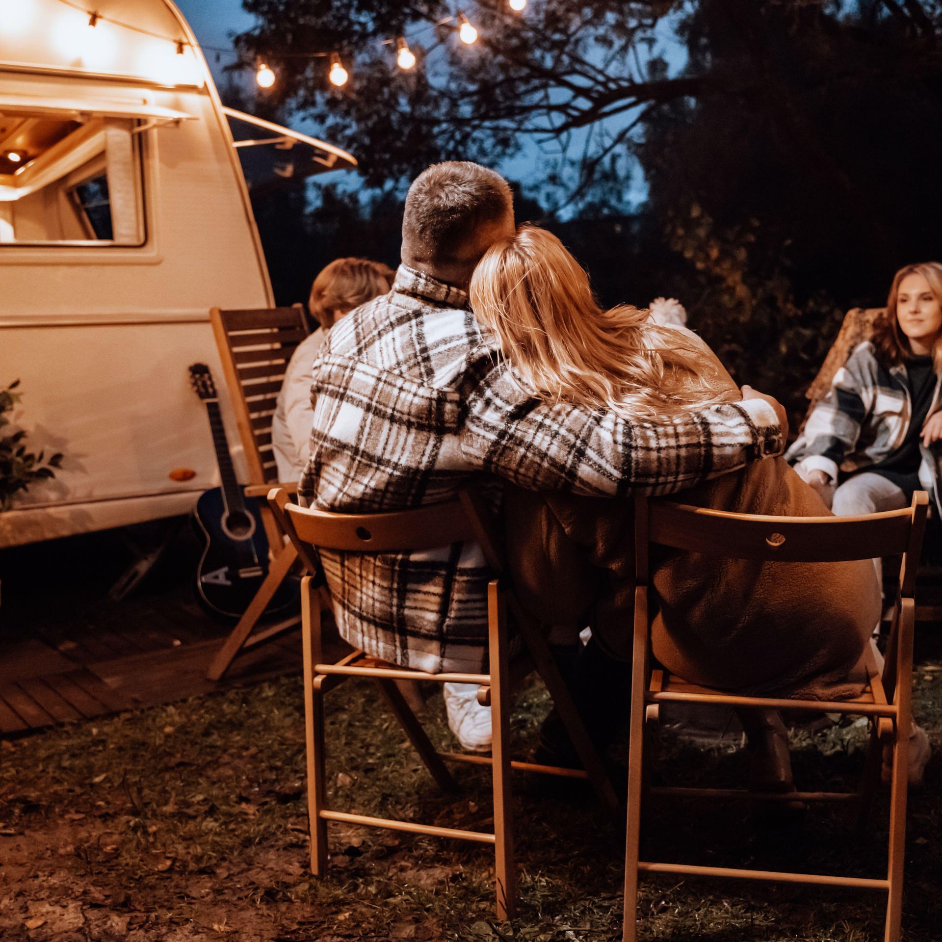 People gather around a campfire, near a camper, under string lights at night.