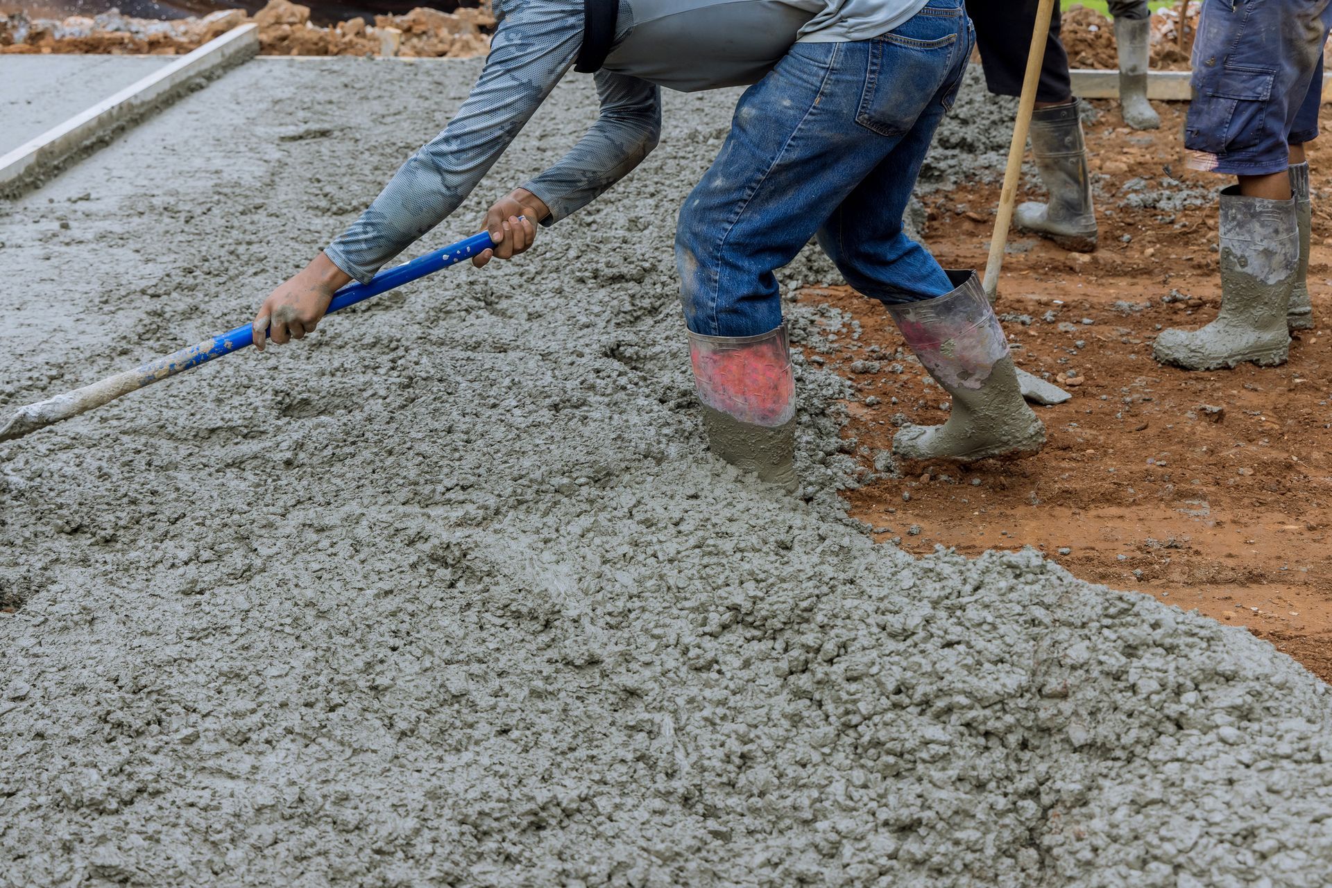 A concrete worker pours wet concrete on the ground, as he flattens it with a stick.