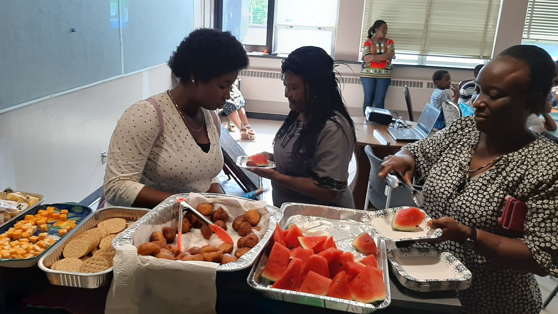 People serving themselves food at a buffet with watermelon, fried dough, and other dishes.