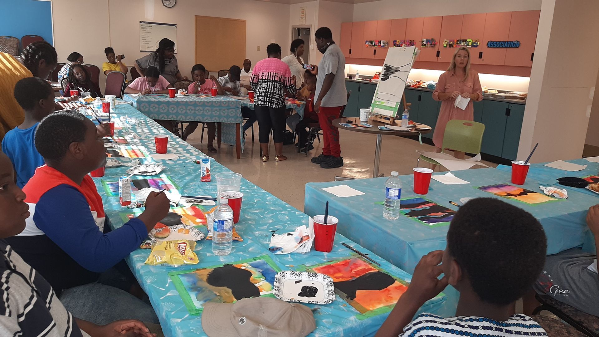 Children and adults in an art class; painting at tables. Instructor speaks near a display; bright room.