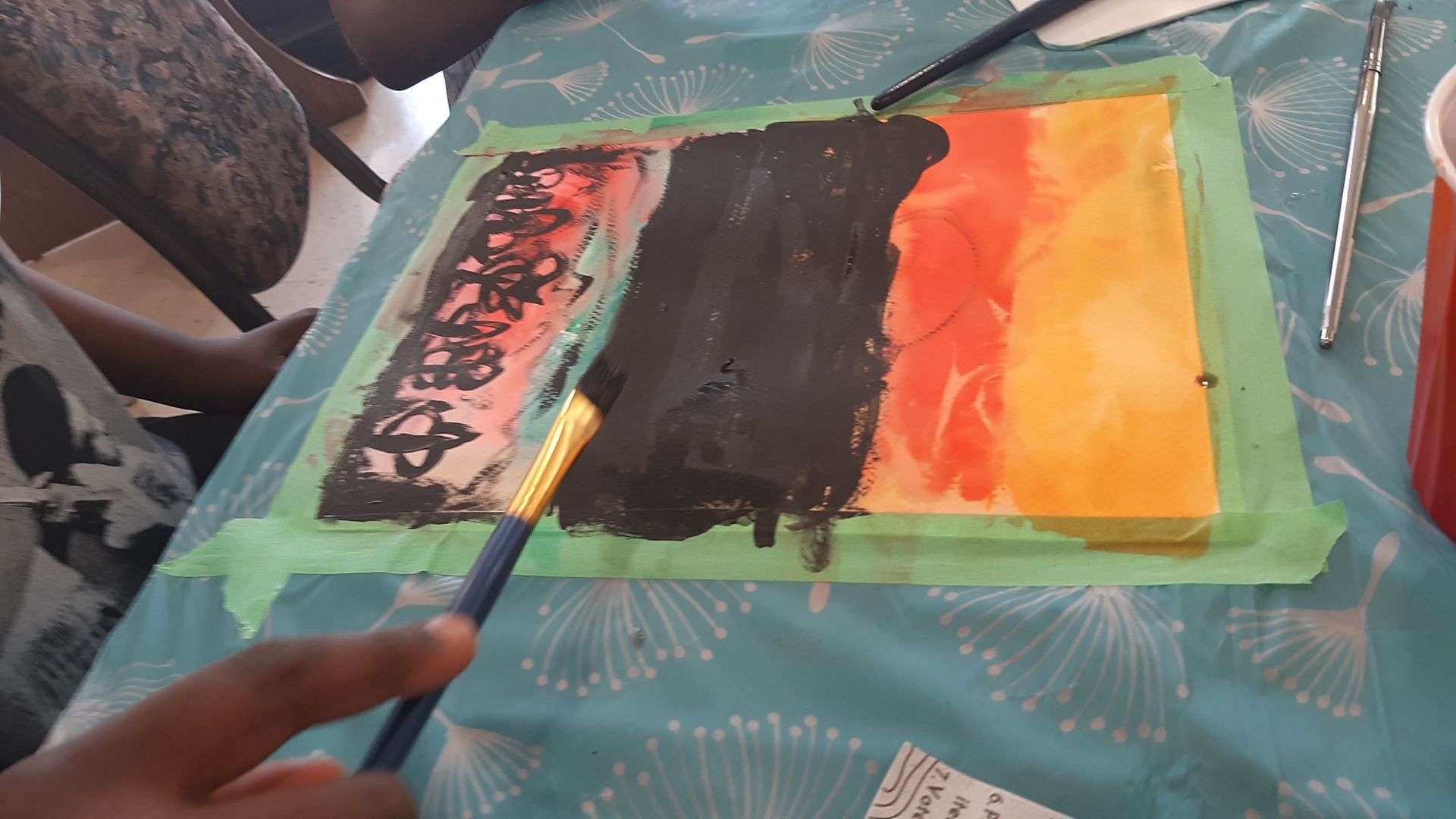 A person painting with black paint onto a colorful artwork, taping on a table.