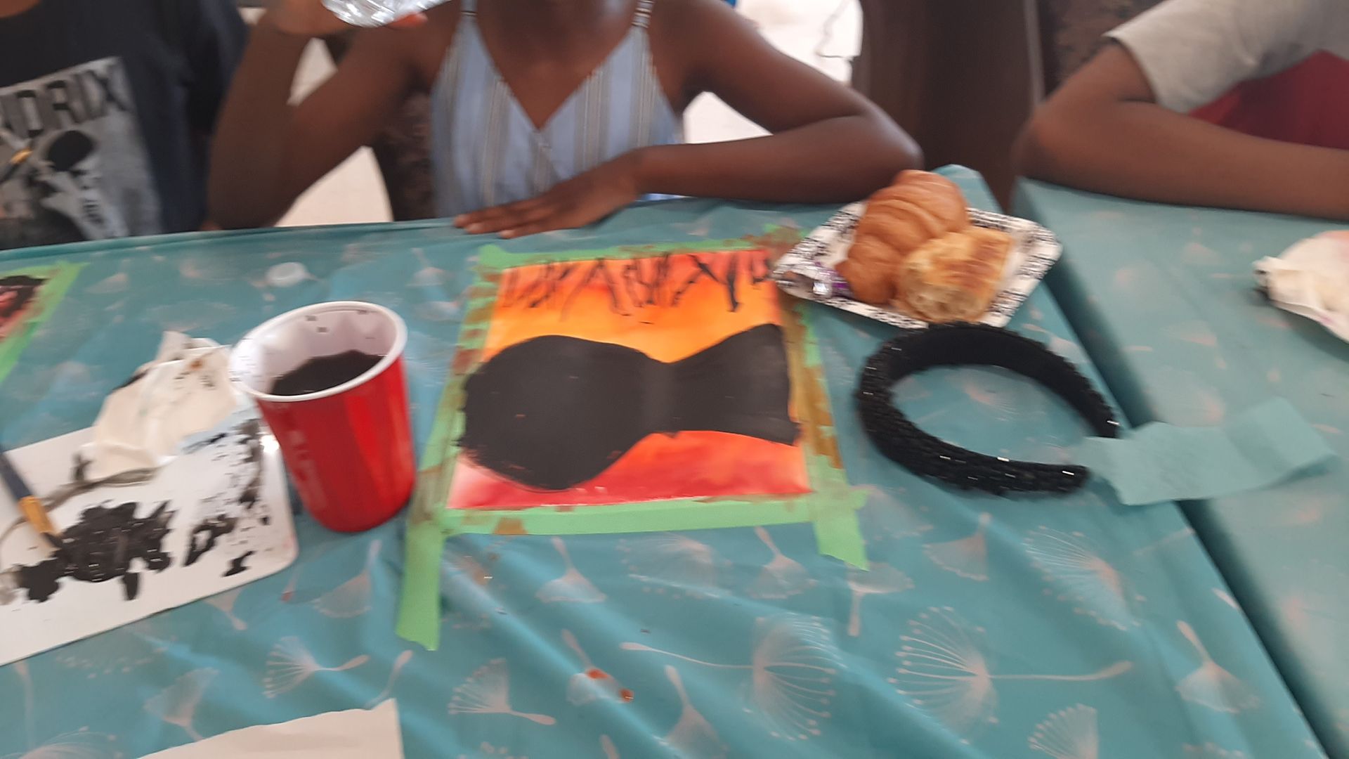 Group of children painting with supplies on a blue tablecloth, including a painting of a guitar, pastries, and a headband.