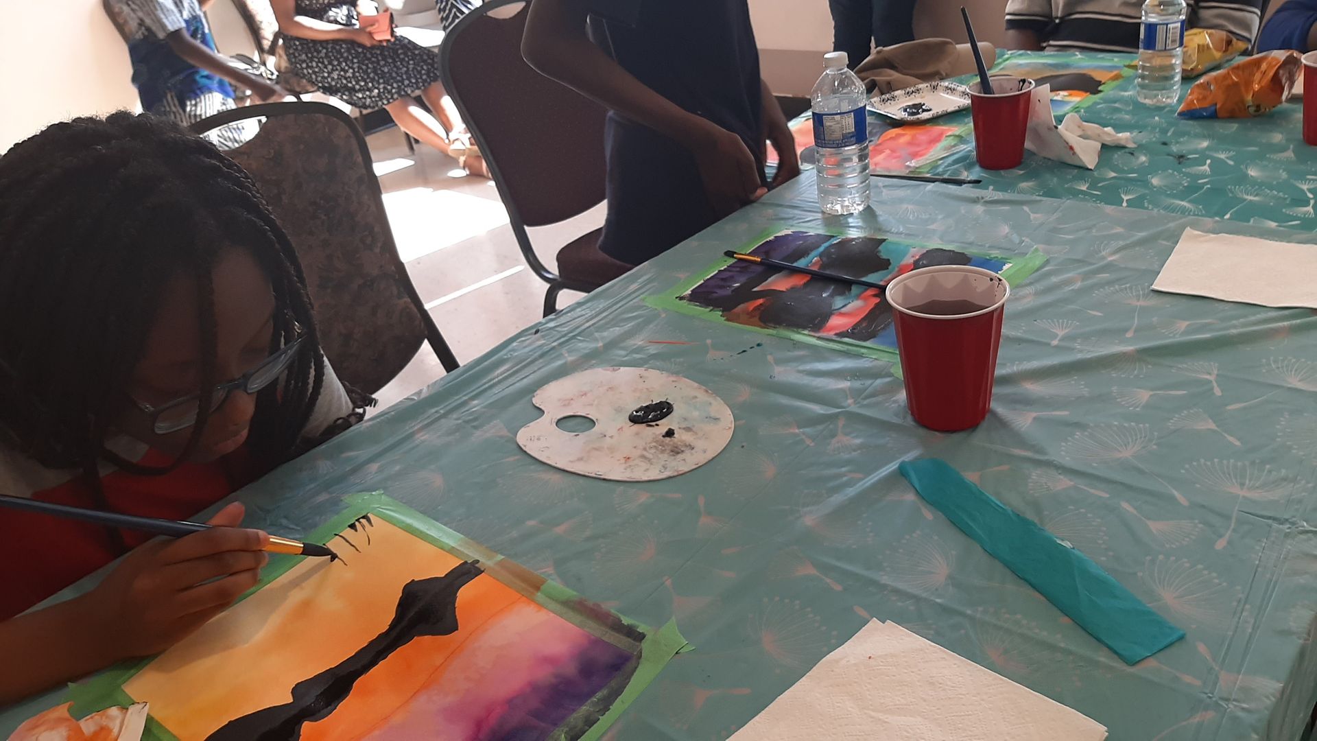 Person painting with watercolors at a table, other people and painting supplies visible.