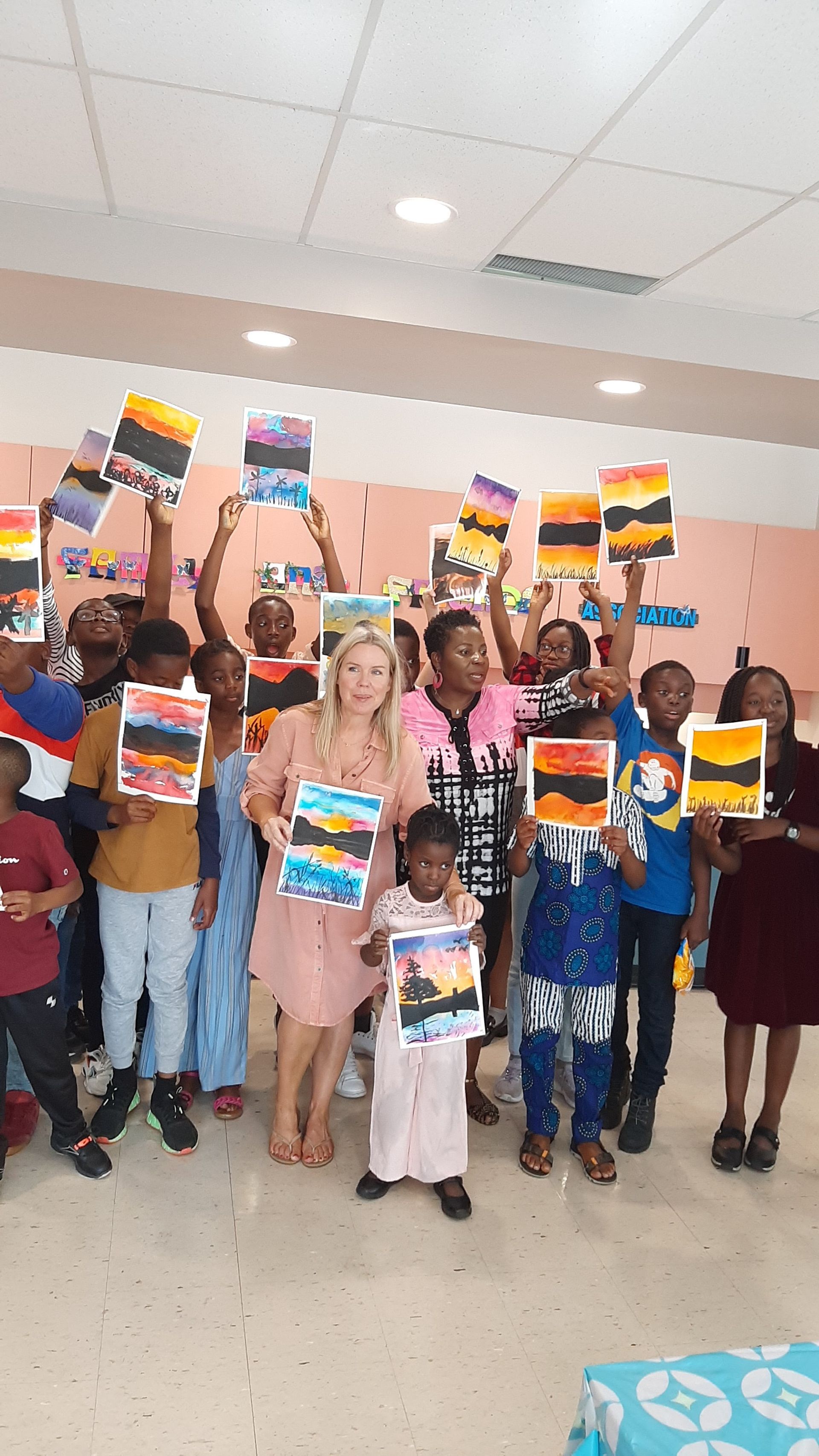 Group of children and an adult holding up paintings of sunsets. Indoors, joyful expressions, vibrant colors.