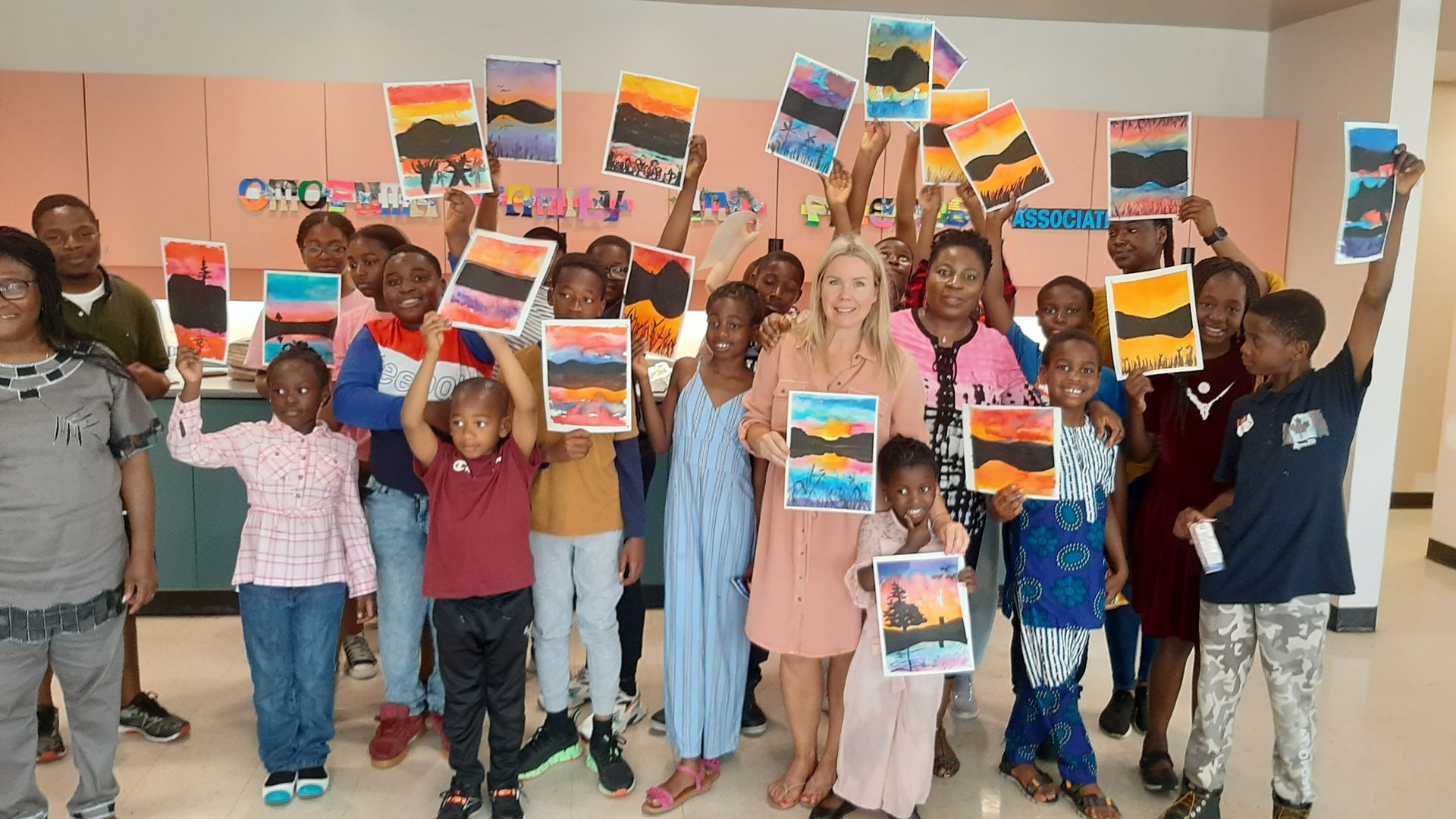 Group of children and an adult holding up landscape paintings, indoors.