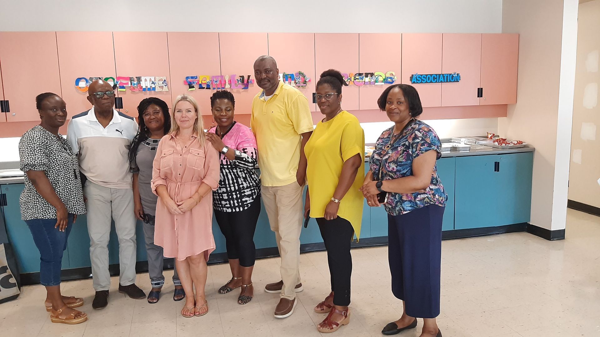 Group of people posing indoors, smiling. Pink, yellow, and blue clothing, light pink cabinets.