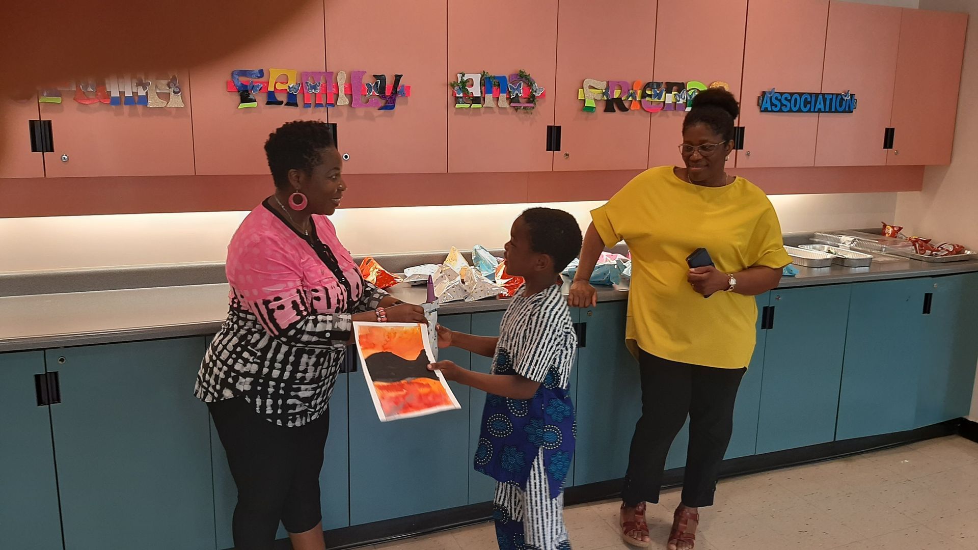 Two women and a child in a classroom. The child presents artwork to one woman.