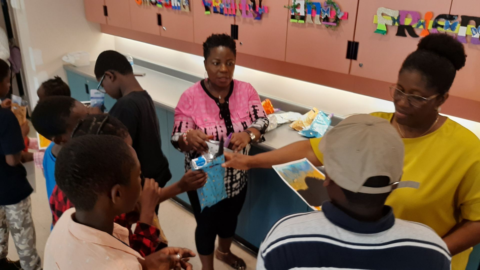 Children and adults in a classroom setting; adults handing out items to the children.