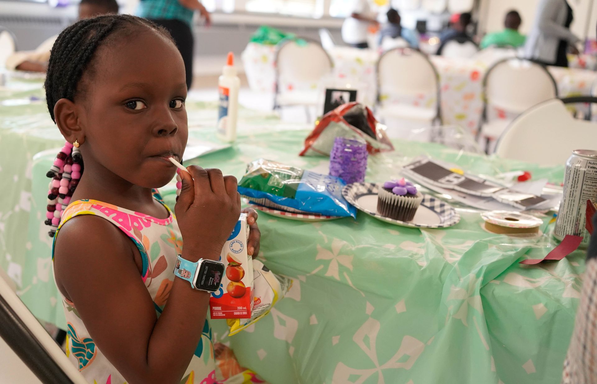 Young Black girl with braids sips a drink at a craft table.
