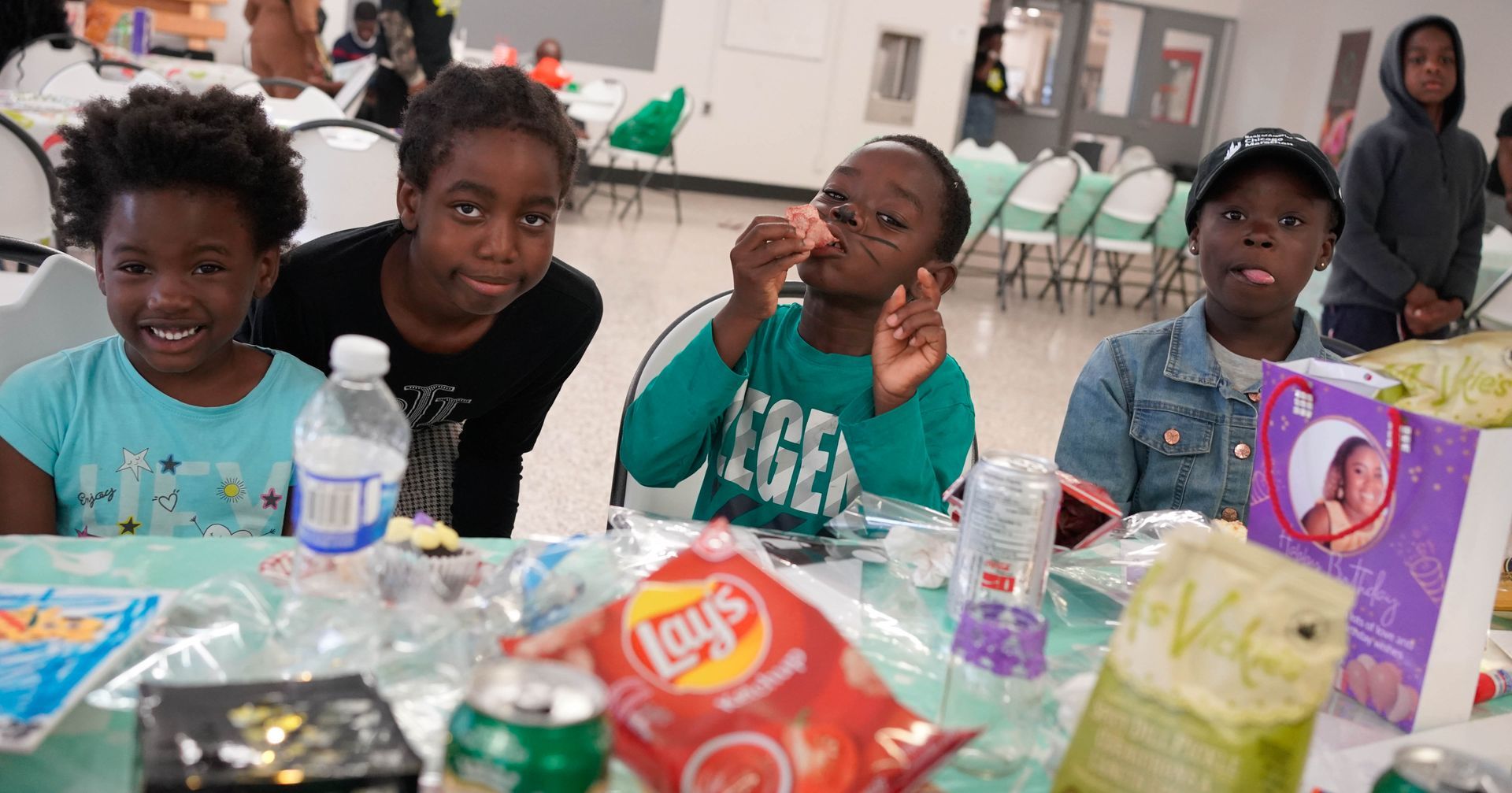 Four Black children at a table, eating snacks, smiling. Indoor setting with green tablecloth and food.