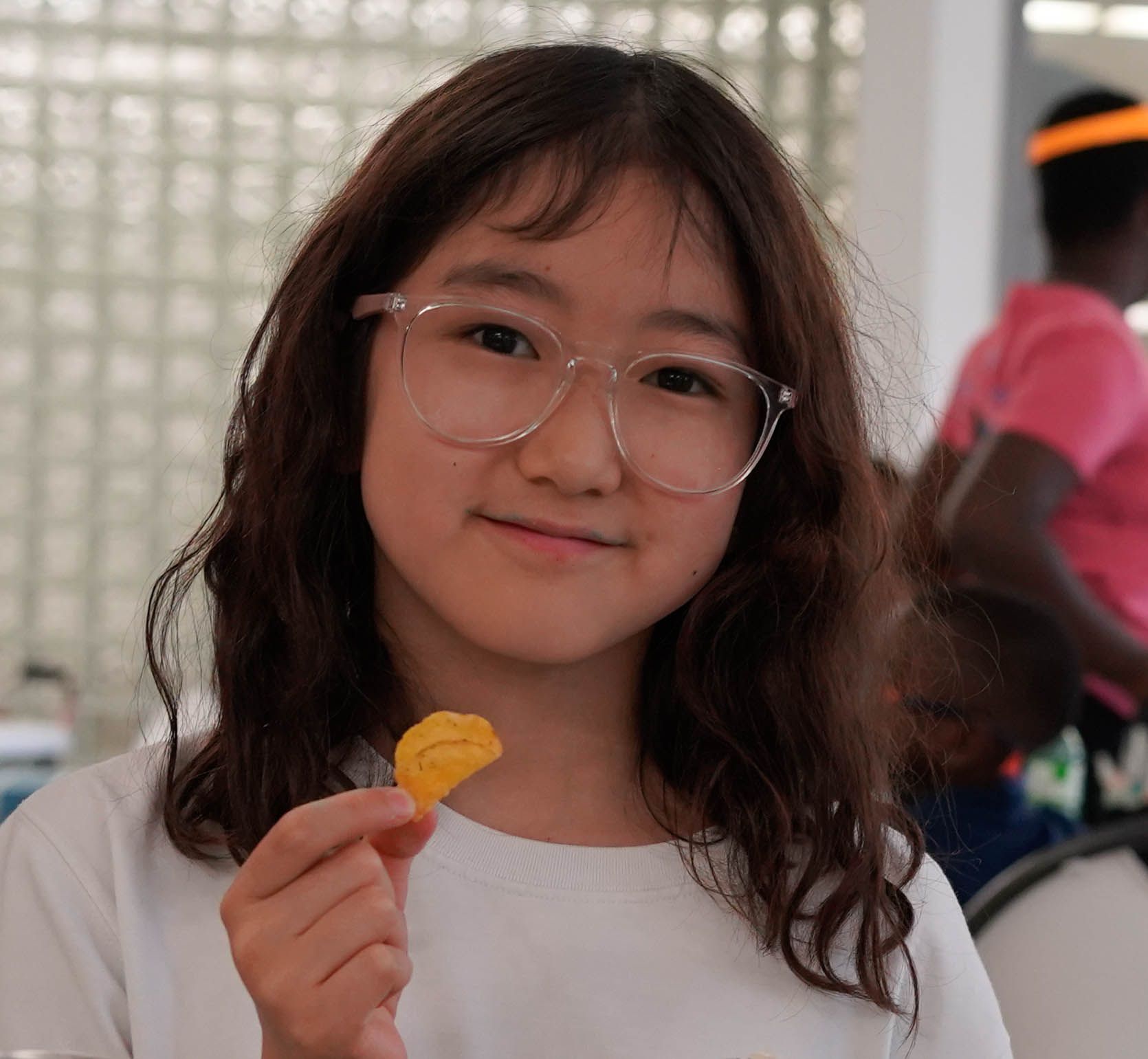 Girl with glasses smiles, holding a potato chip; blurred background with people.