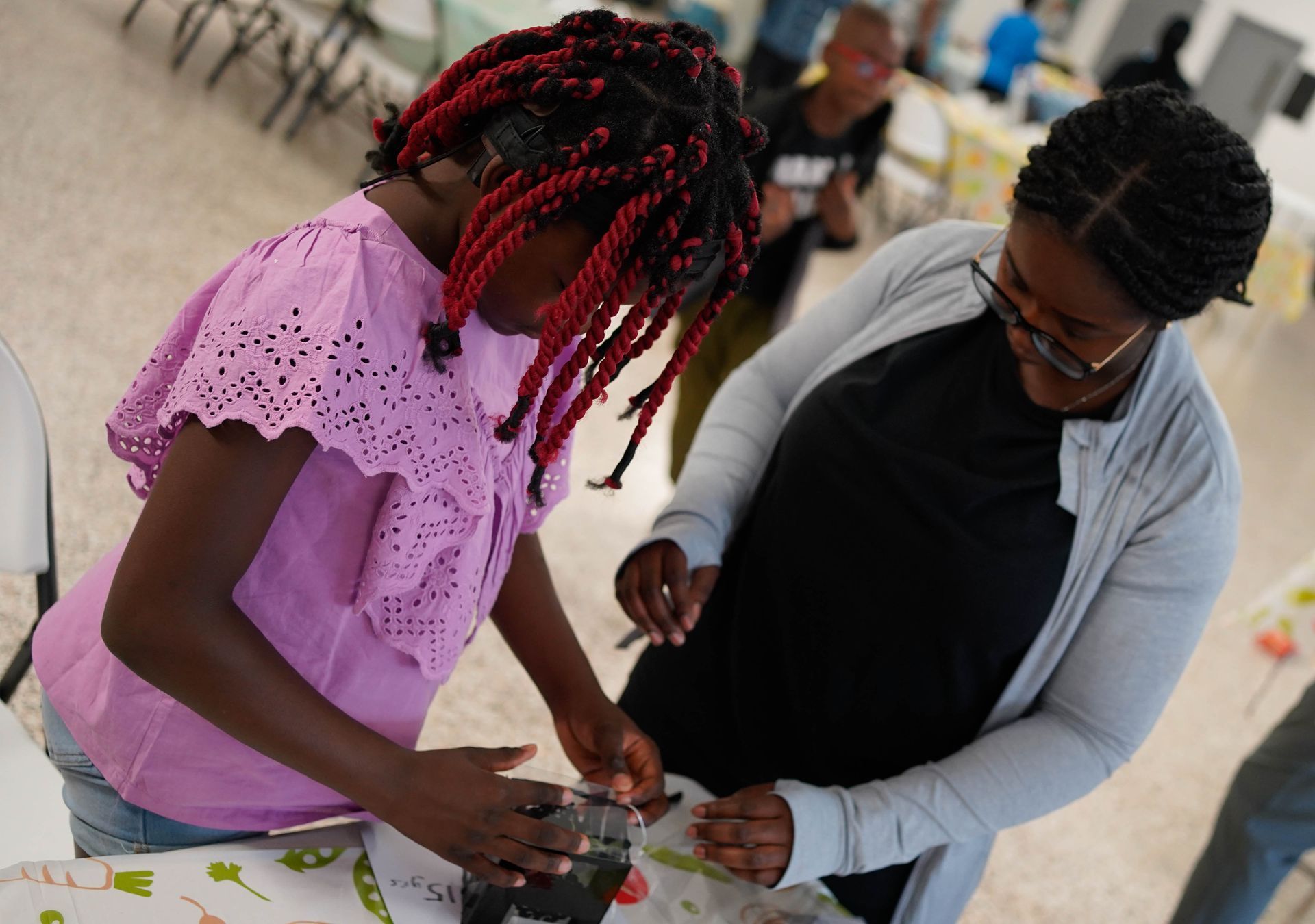 Two Black women lean over a table in a room. One braids her hair and wears a pink shirt. The other, smiles.