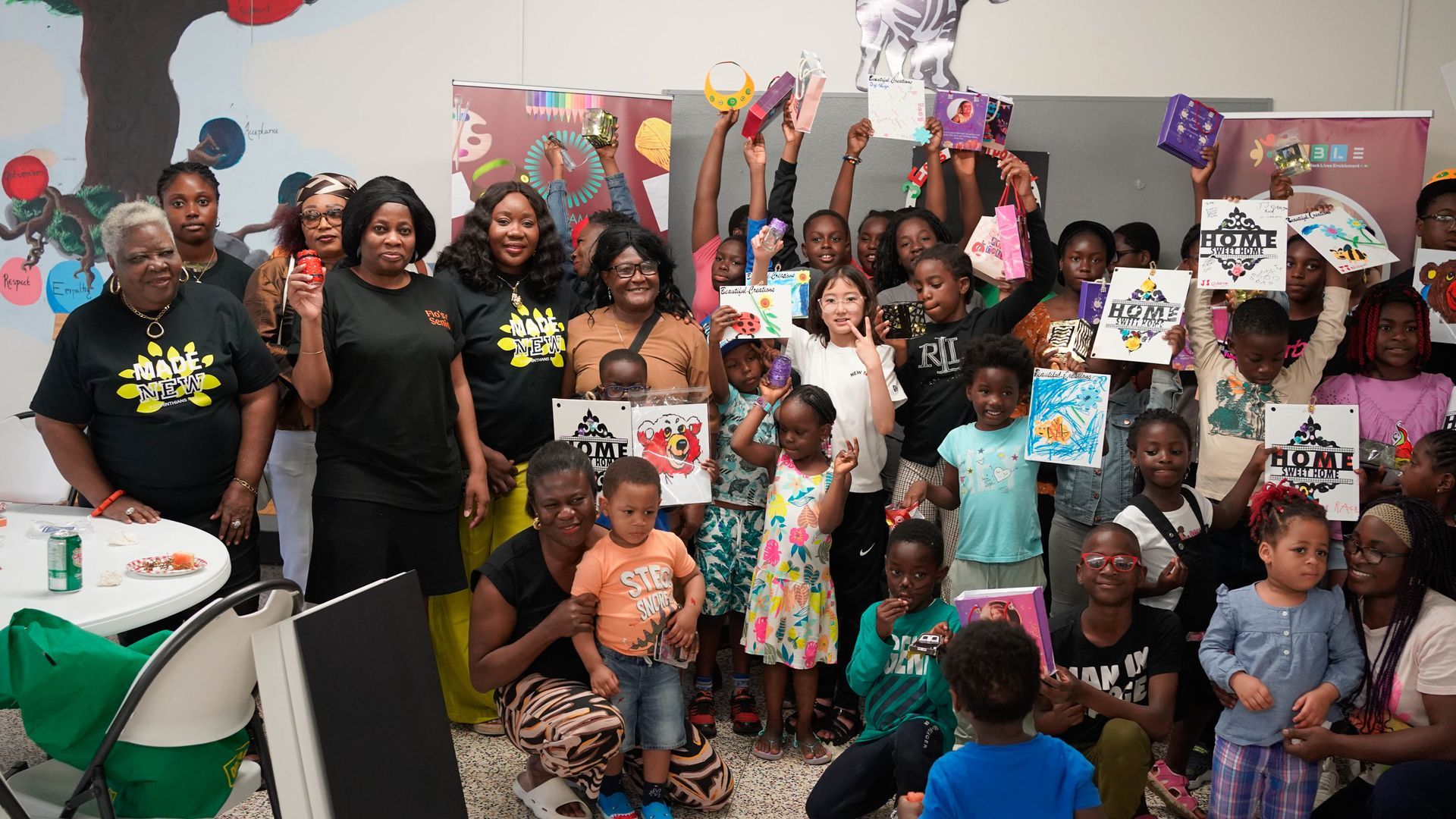 Group of people, children and adults, hold up artwork, smiling. Indoors, bright colors.