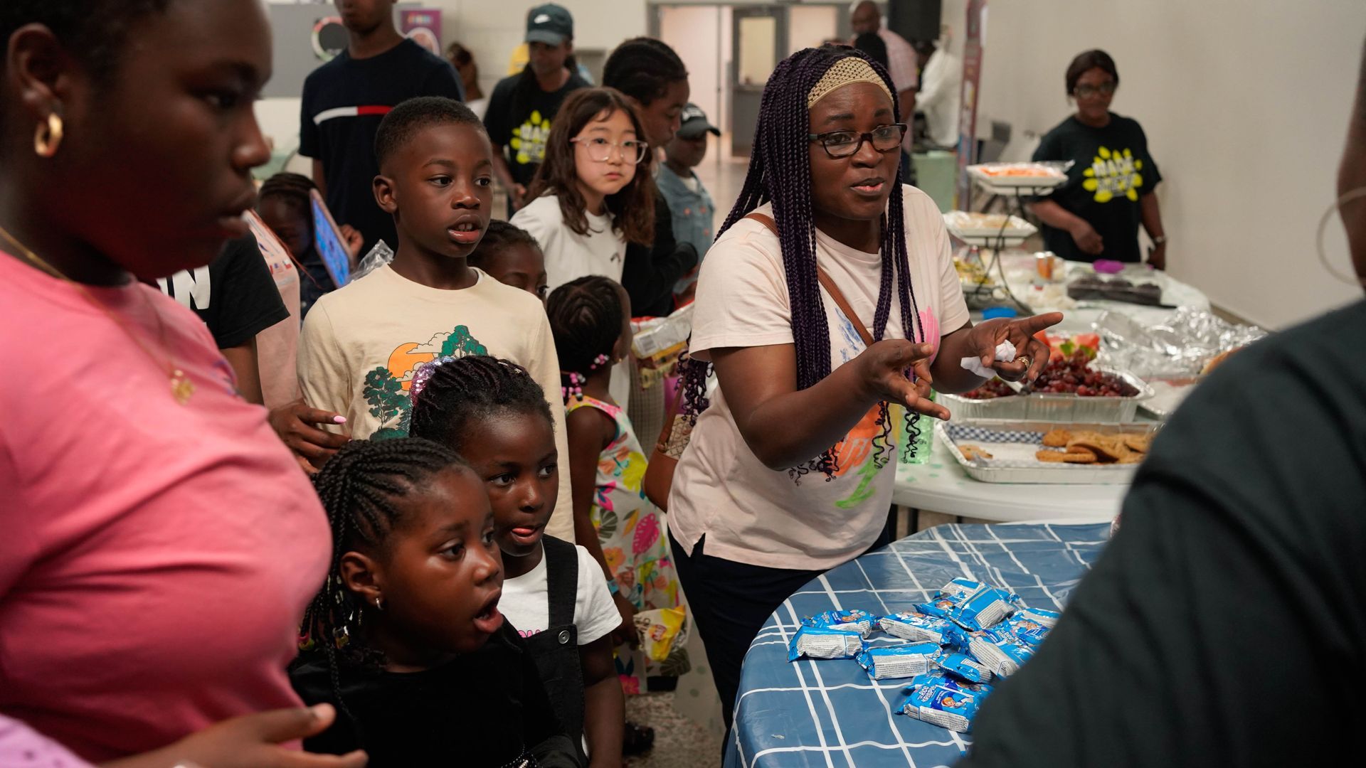 People at a community gathering, some looking at food on a table. Many Black children and adults in a room.