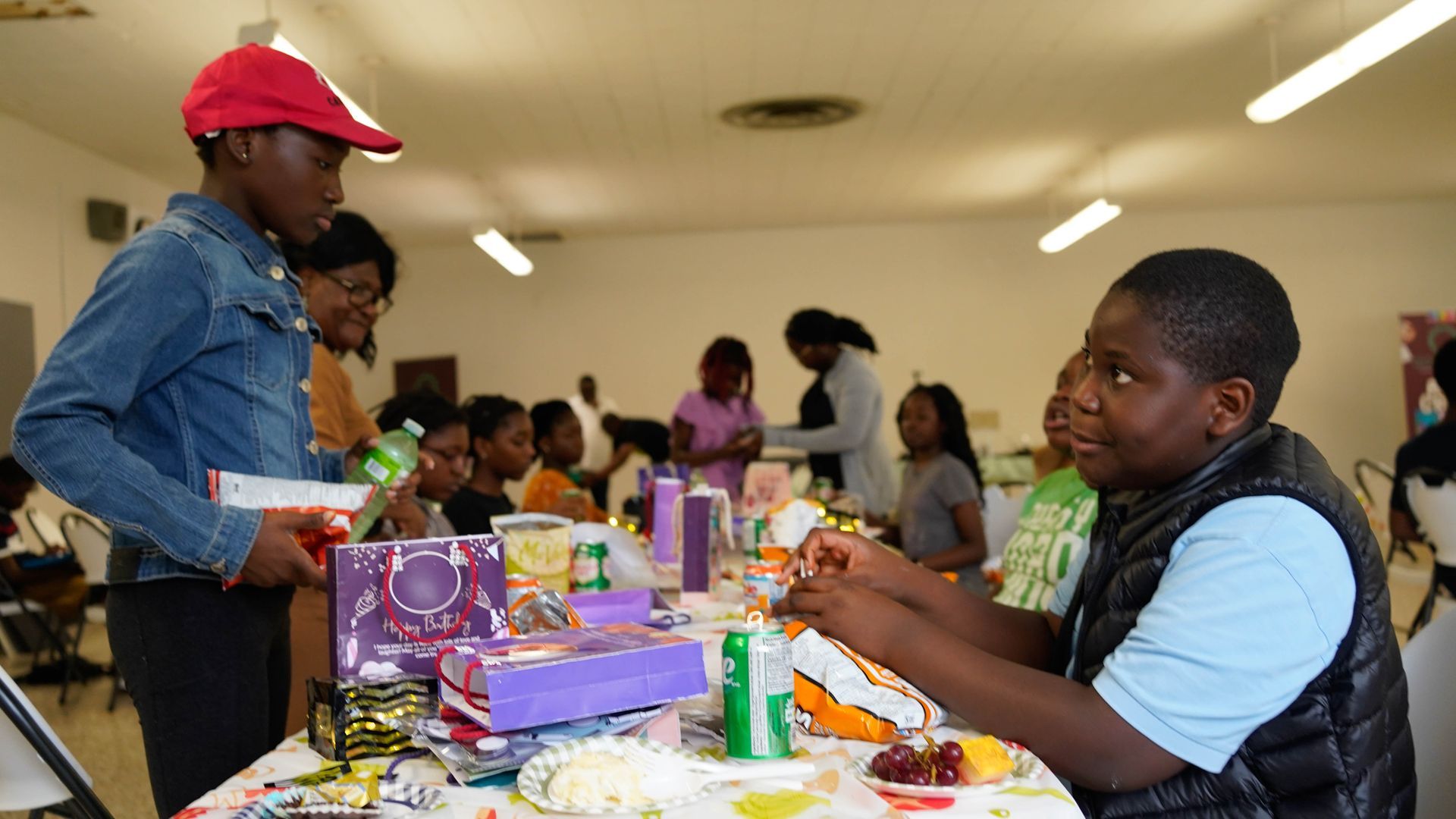 Children at a party, one boy in a vest opens a snack, another in a red cap offers a drink.