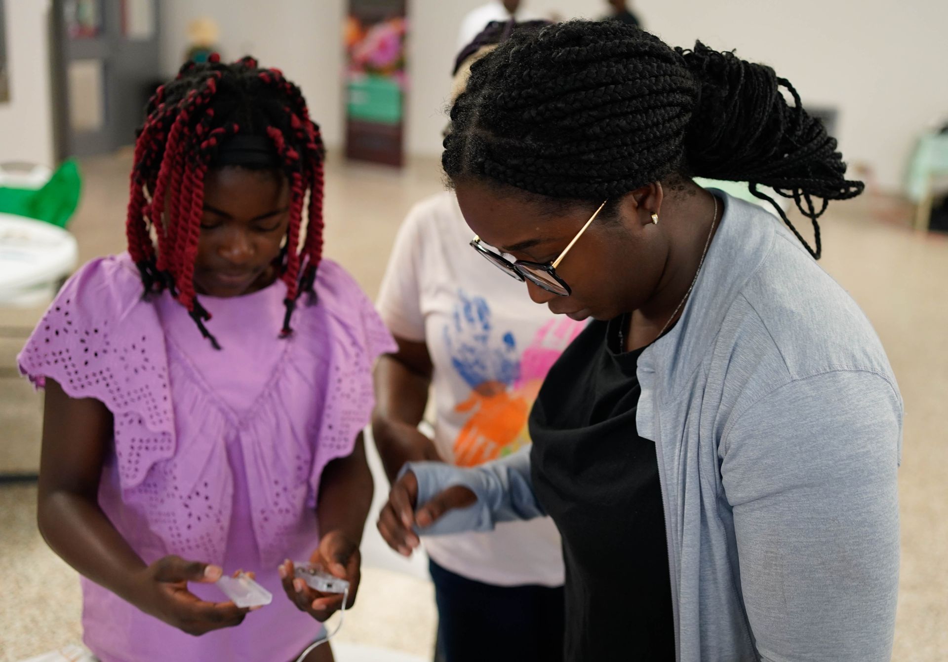 Two young Black women examining an object, looking down intently, in a room with another person standing behind them.