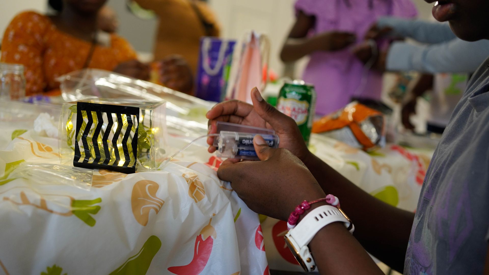 Person holding small container at a table with items for a craft, with others in background.