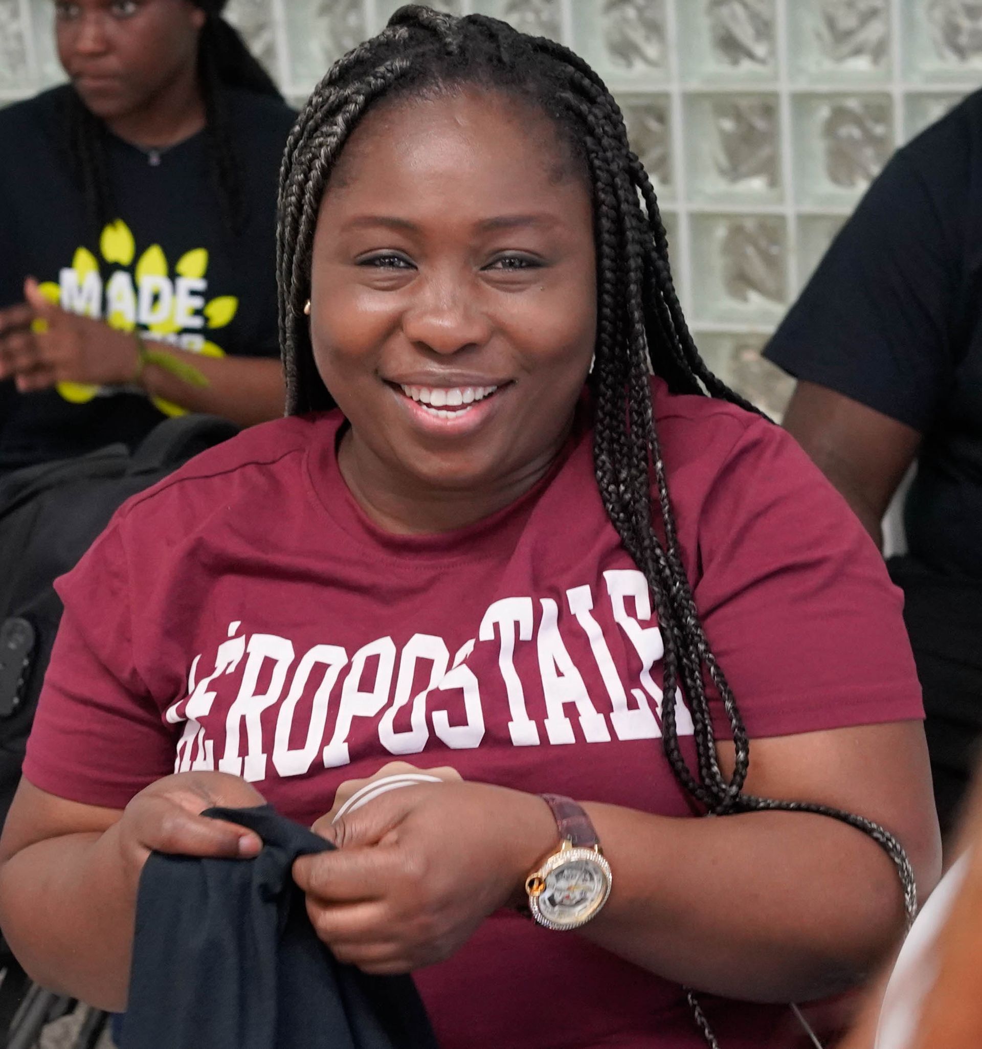 Woman with braided hair smiles, wearing maroon Aeropostale shirt and watch, in a room with others.