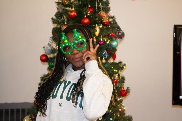 Person wearing festive glasses, giving the peace sign, stands in front of a decorated Christmas tree.