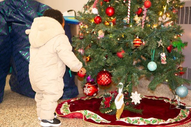 Child in snowsuit reaching toward decorated Christmas tree.