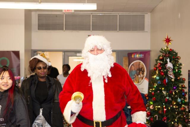 Santa Claus in red suit, walking in a room with a decorated Christmas tree and people.