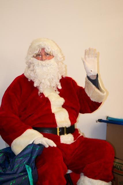 Santa Claus waving, sitting in a room, wearing a red suit with white gloves and beard.
