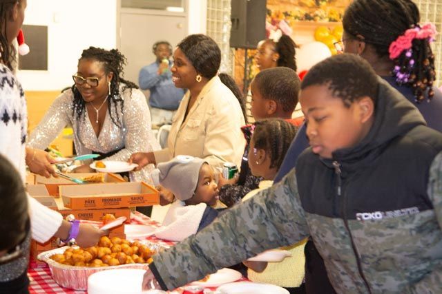 People at a community event, choosing food from a buffet table. Children are present, festive mood.