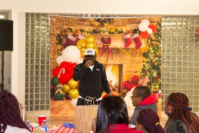 Man speaking at a Christmas event with balloons, fireplace backdrop, and audience in a room.