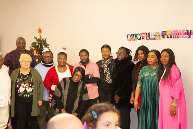 Group of people smiling at a Christmas gathering, against a white wall with colorful text and a small tree.