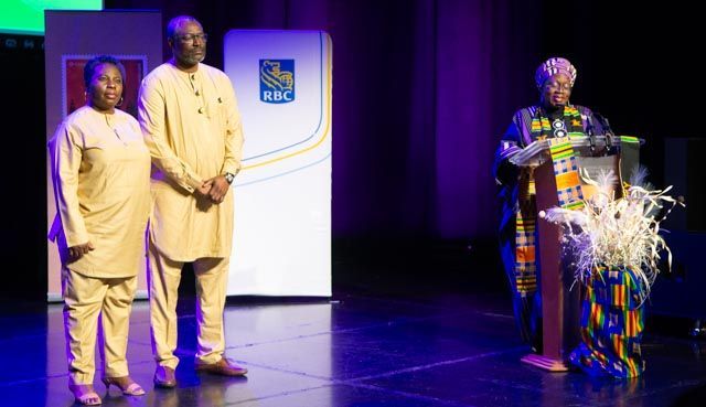 Three people on a stage; two in yellow clothing, one speaking at a podium in colorful attire.