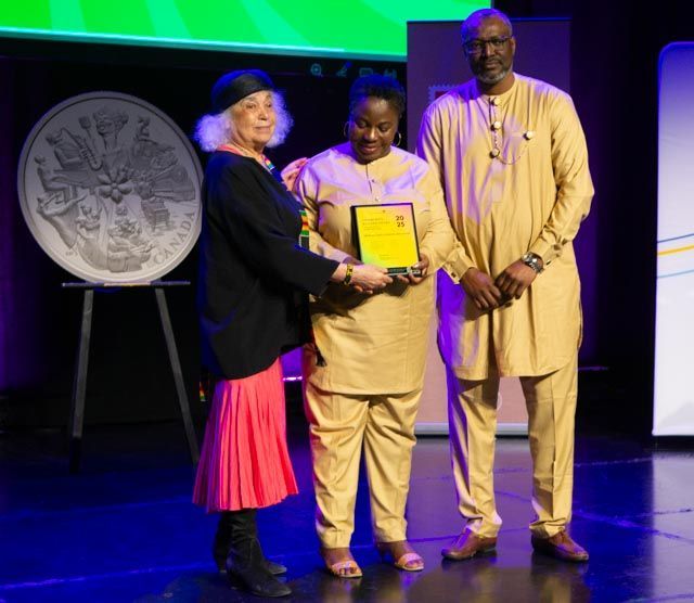Woman in pink and black outfit presenting award to woman in yellow suit, man in yellow suit stands next to them.