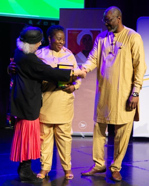 Three people on stage: woman in black embraces woman in yellow suit as man in yellow suit shakes hands.