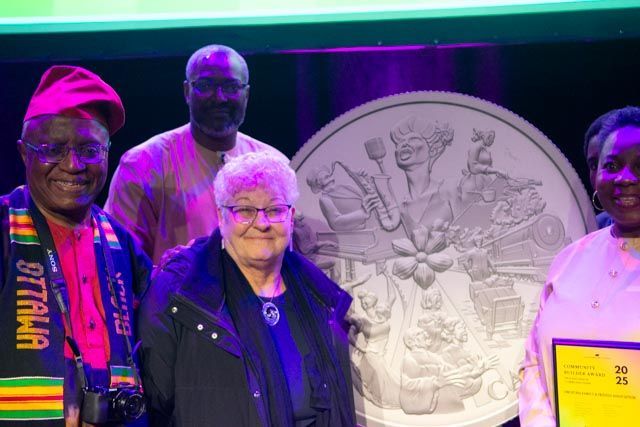 Group of people pose with a large commemorative coin, likely an award presentation at a theater or auditorium.