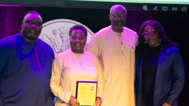 Four people pose onstage, one holding a certificate. Background includes a logo and purple lighting.