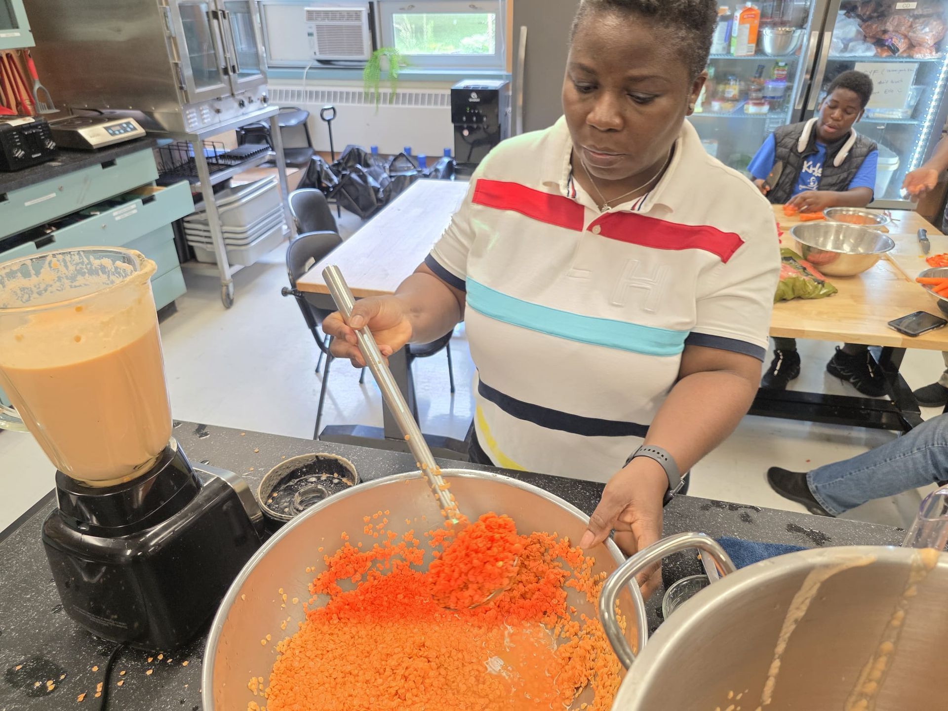 Woman stirring food in a large metal bowl, blender to the left. Other people in the background.