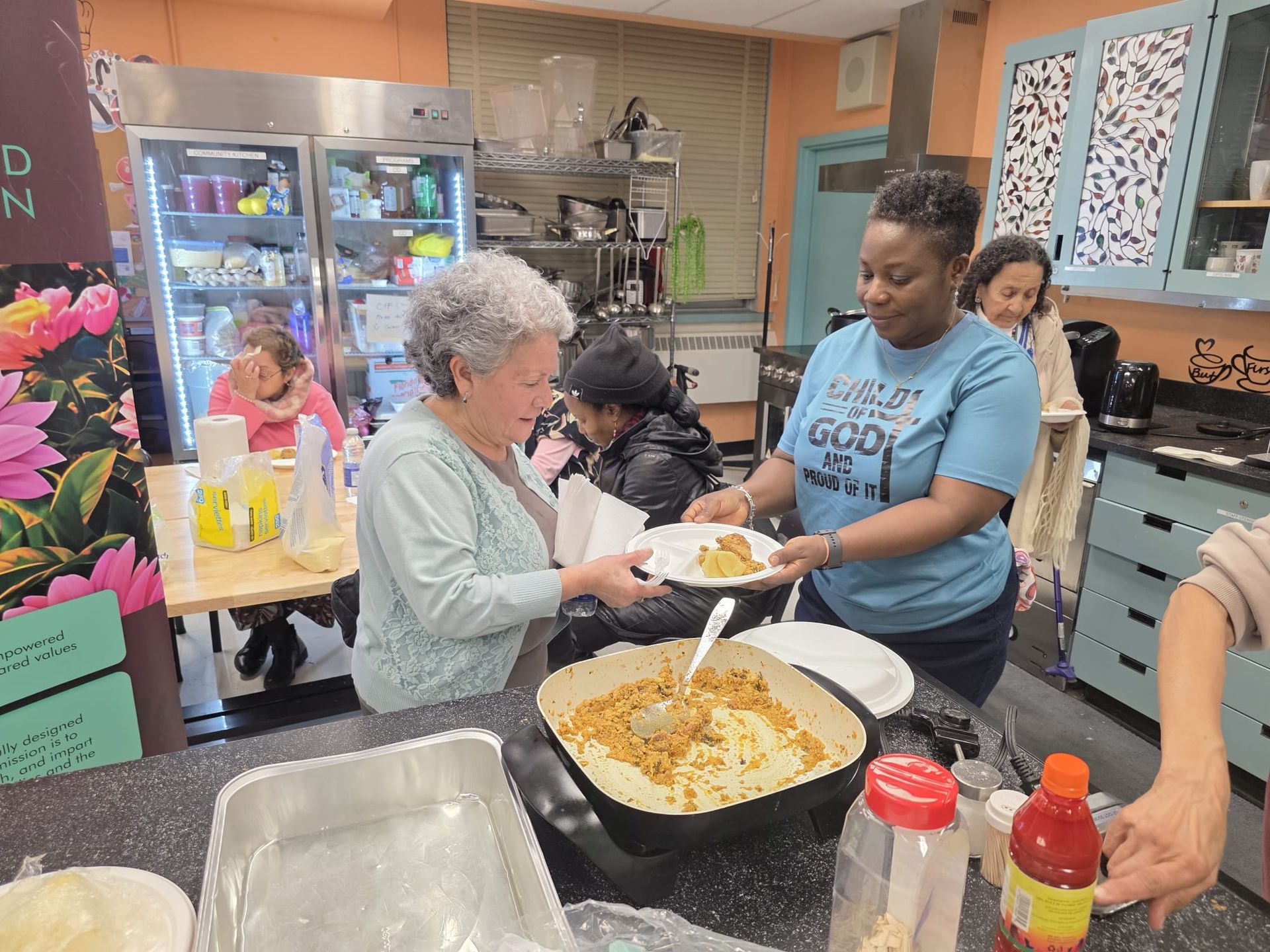 Woman serving food to another woman from a large pot in a kitchen. Other people nearby.