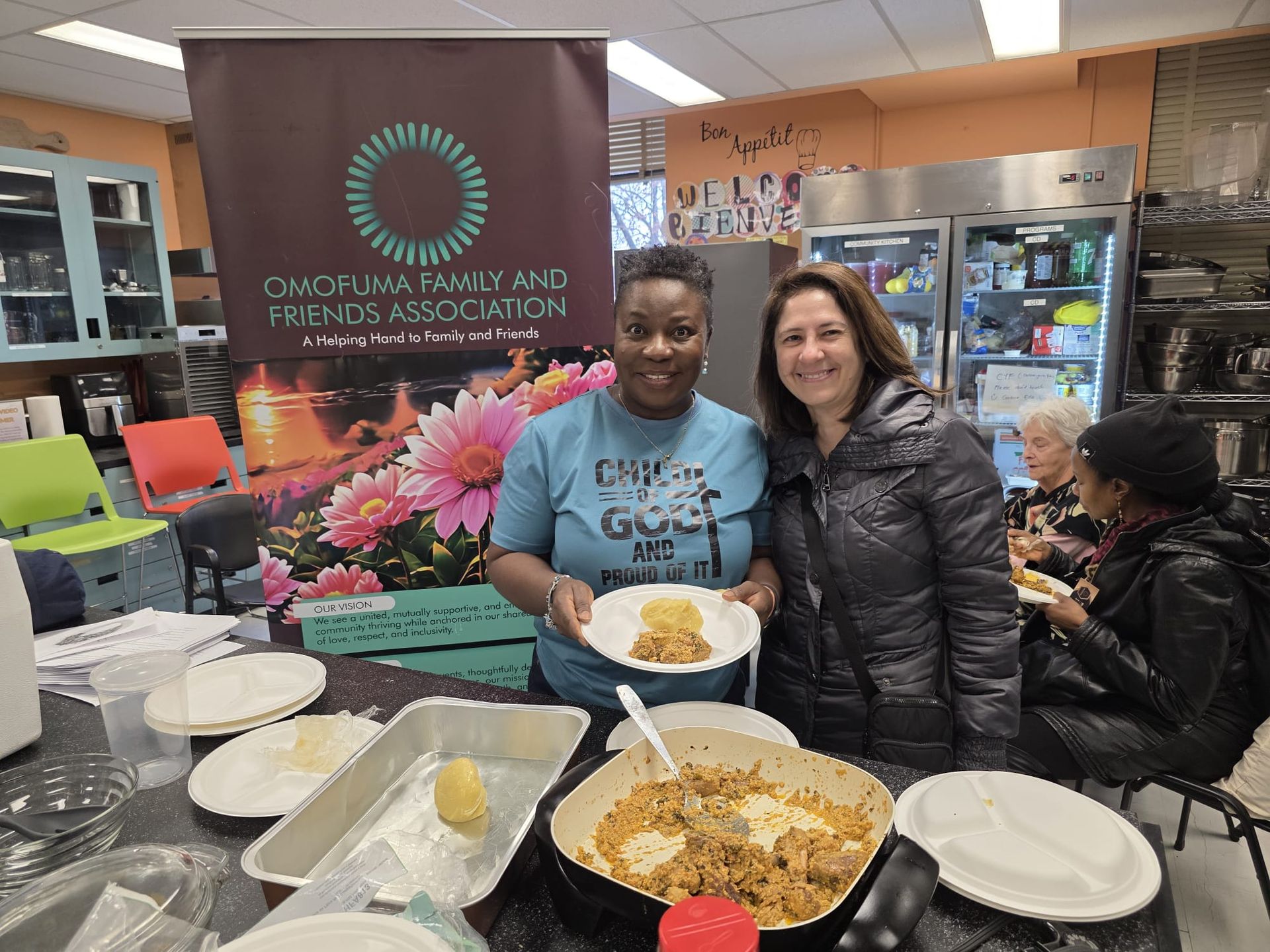 Two women holding plates of food, smiling in a kitchen with a banner for an association.