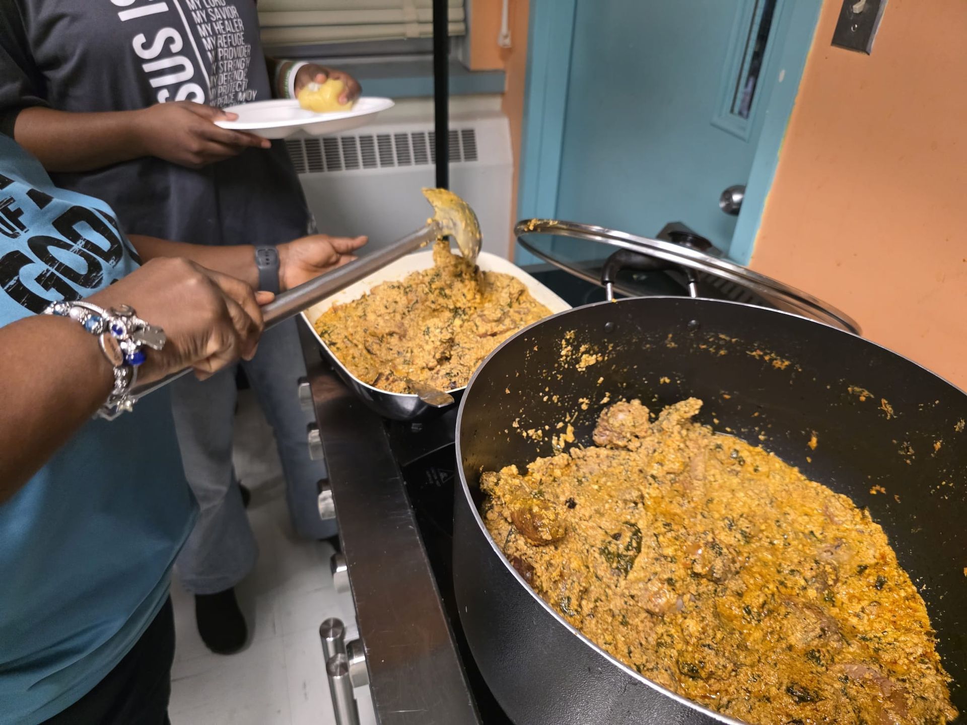 Person ladling food from a pot onto a serving dish. Another person holds a plate. Kitchen setting.