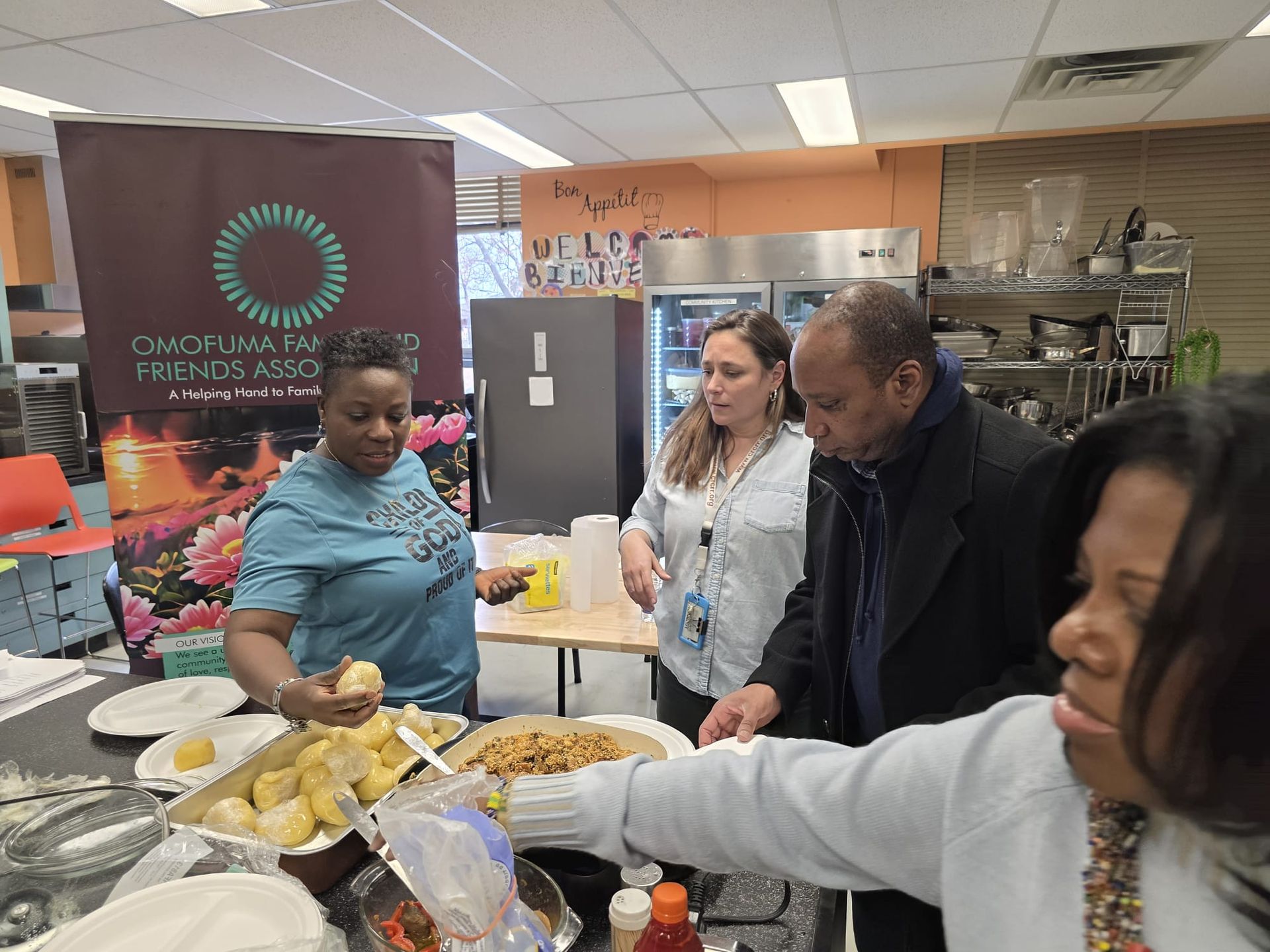 People serving food at a gathering near a banner. A kitchen and dining area are visible.