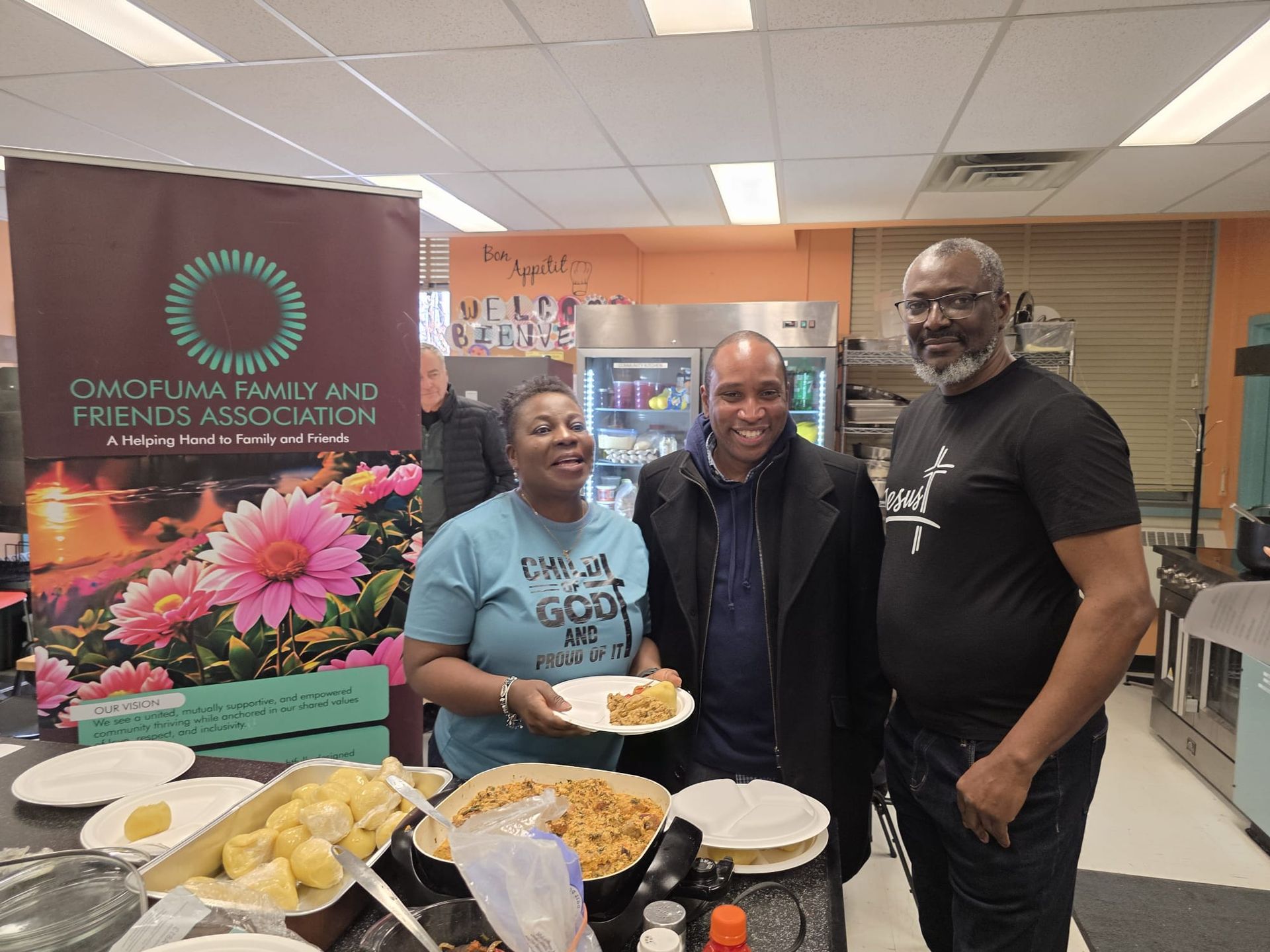 Three people smile at a food table, with a banner behind them. Woman with food, man, and man.