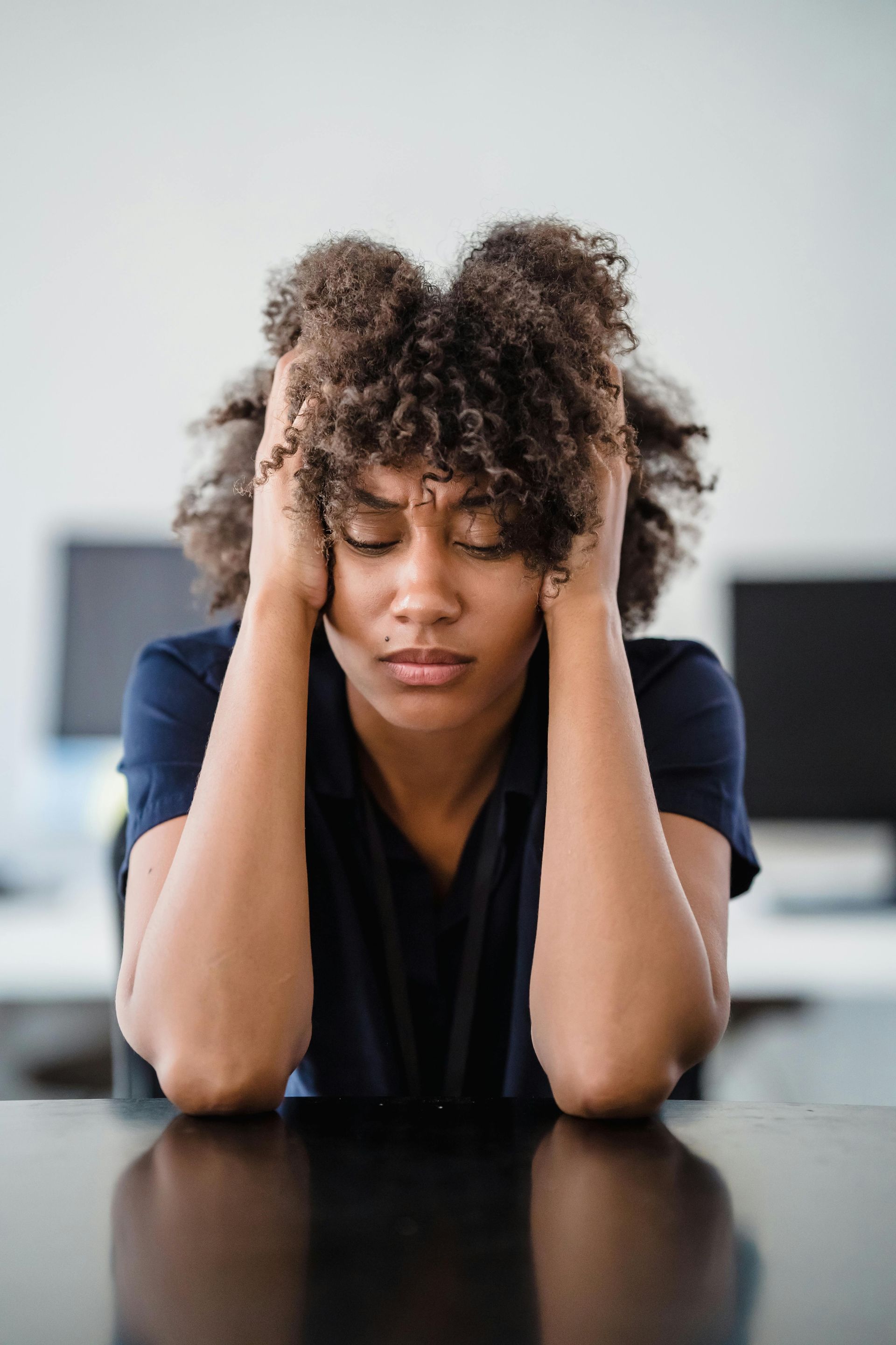 Overwhelmed mom sitting at desk with eyes closed, holding head in stress and exhaustion.