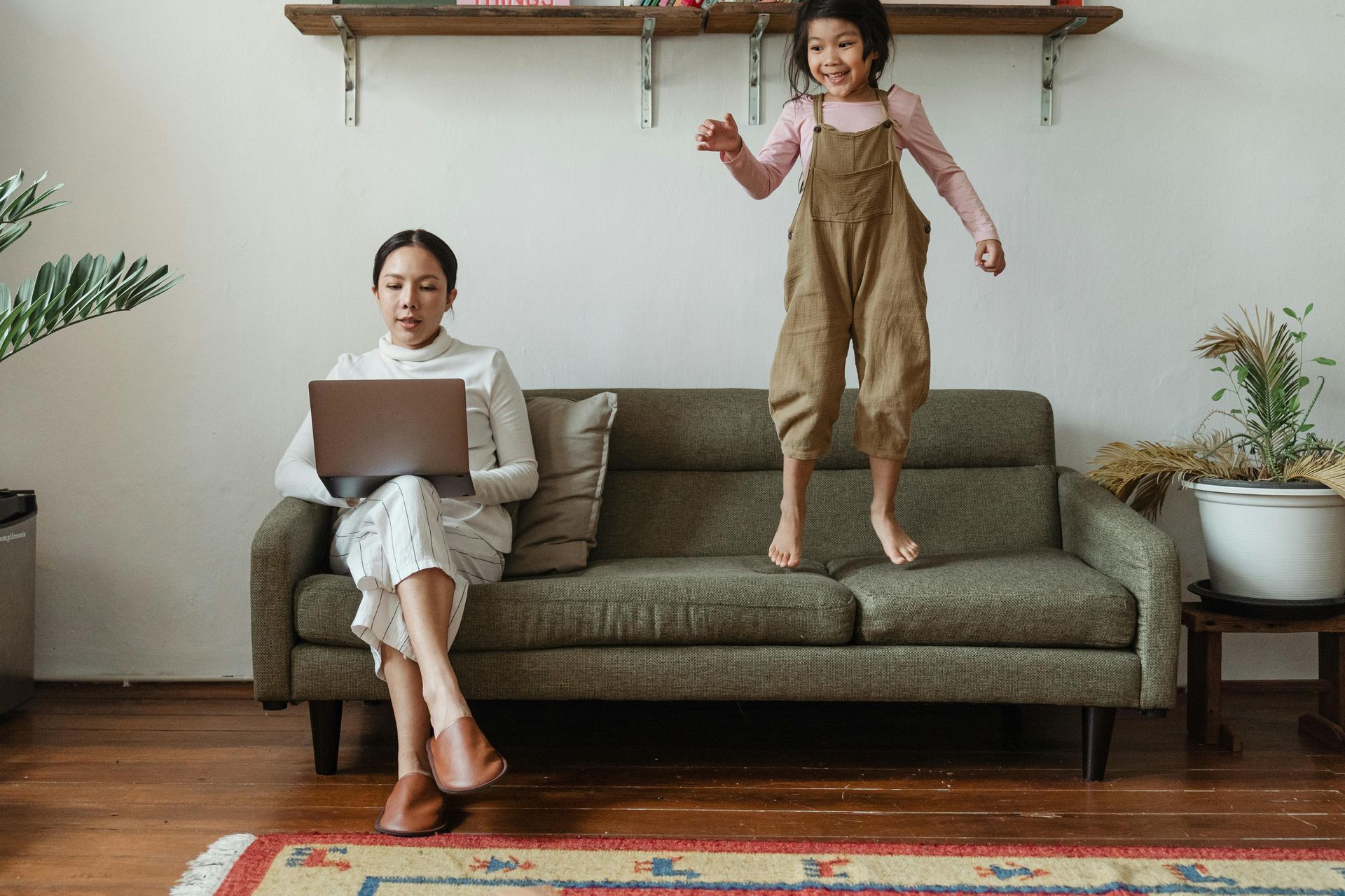 Burned out mother on computer sits to regulate while child jumps on couch