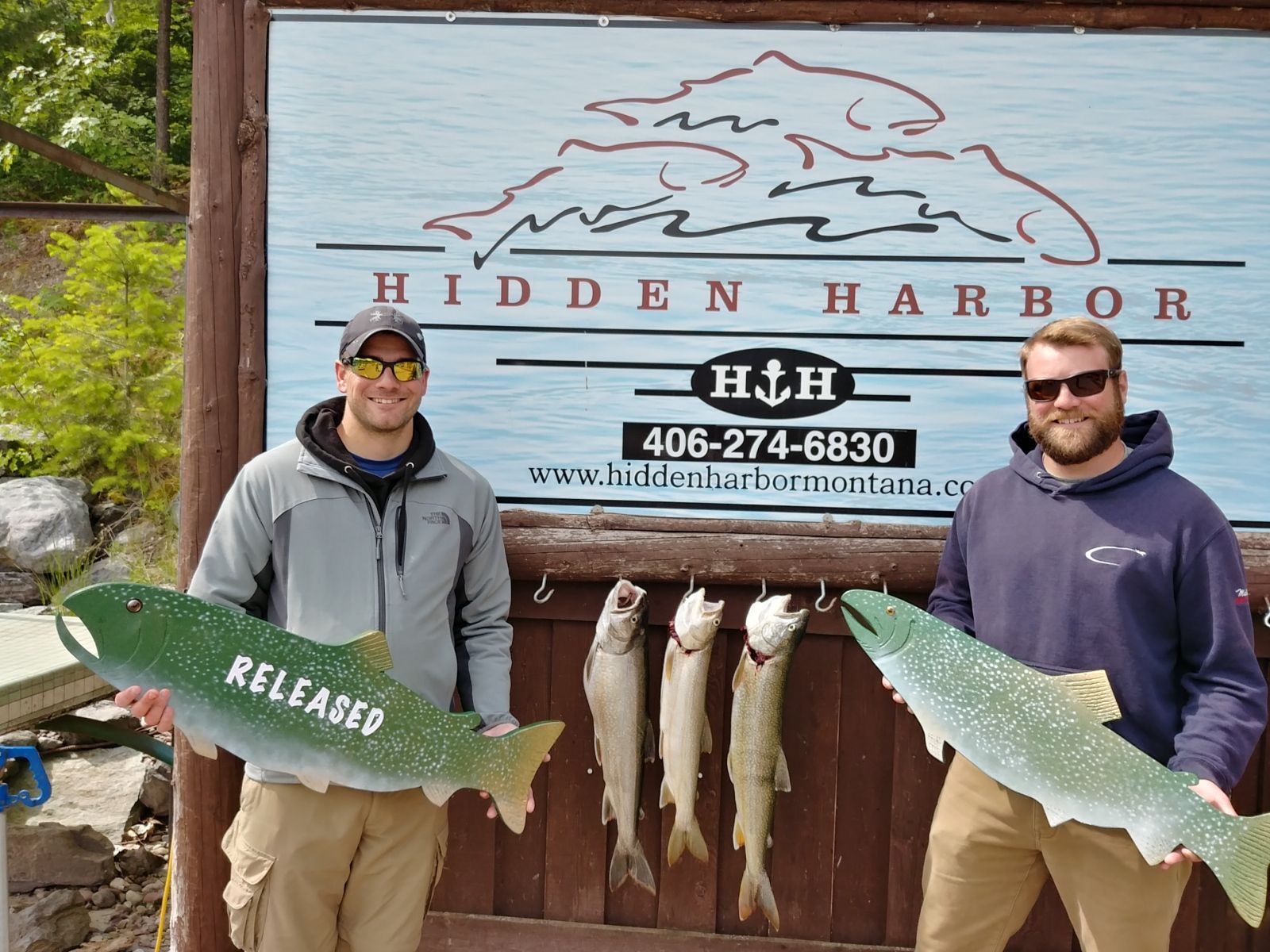 Two men are holding fish in front of a sign that says hidden harbor
