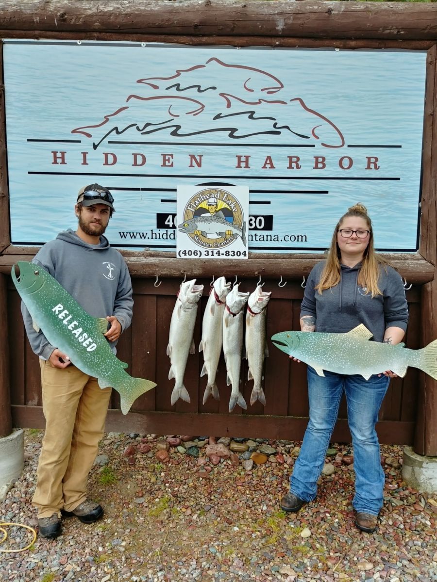 A man and a woman are holding large fish in front of a sign.