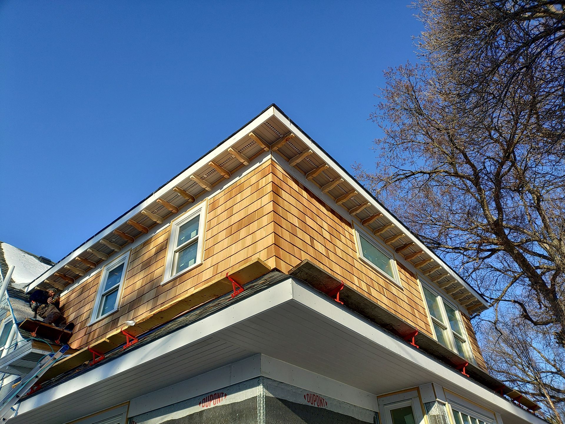 House corner under construction with wood shingle siding and a white trim, against a blue sky.