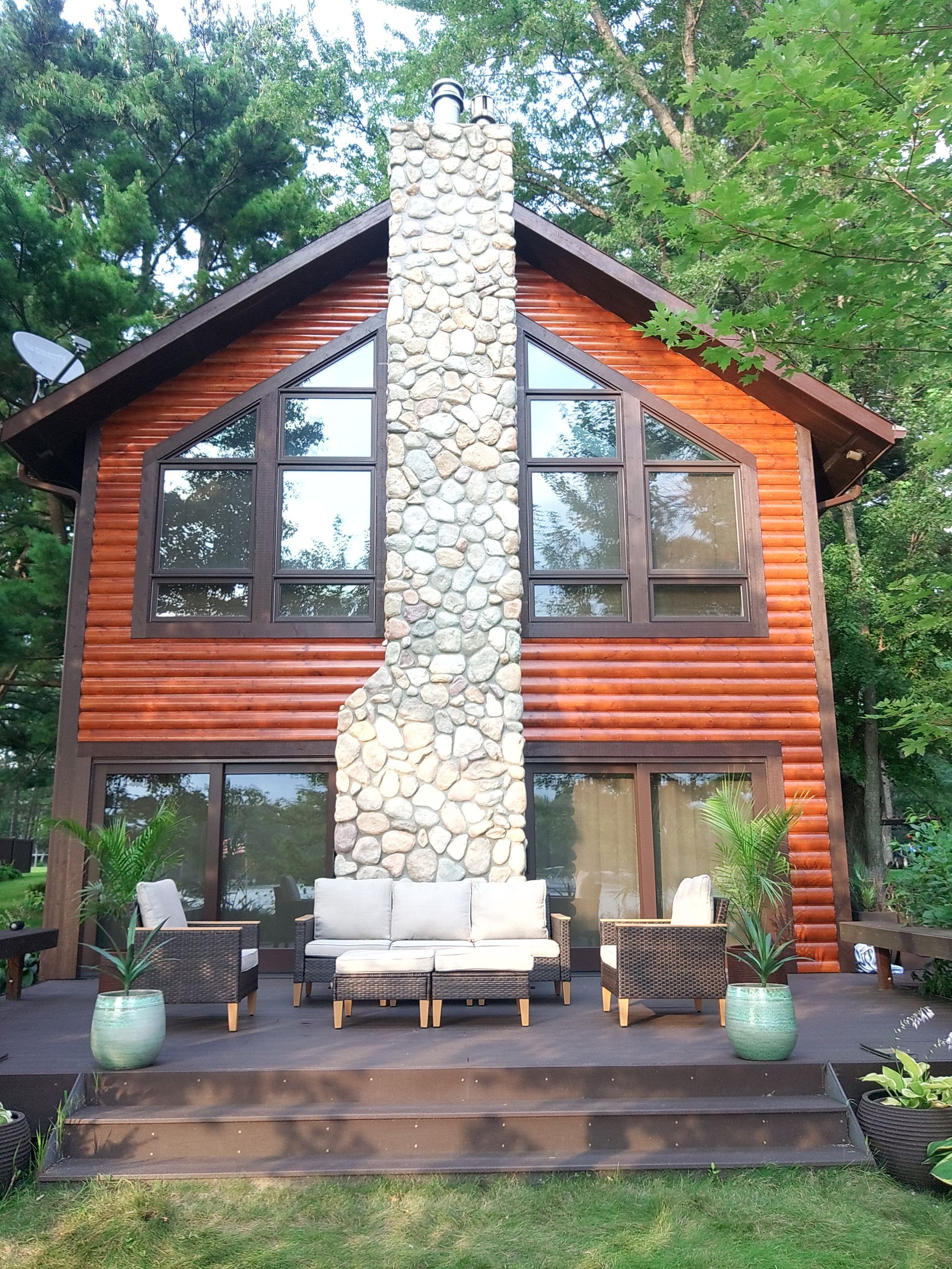 Log cabin with stone chimney, large windows, and a deck with seating, surrounded by trees.