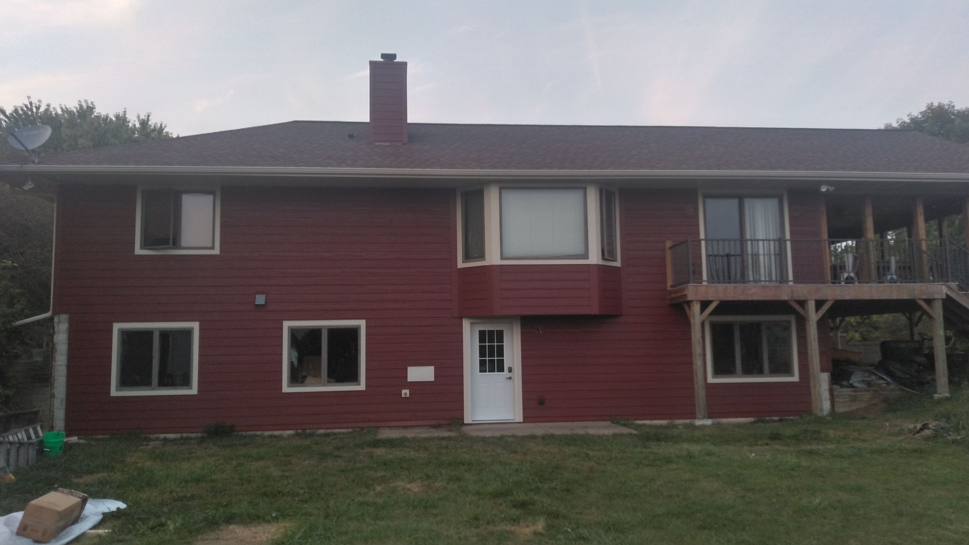 Red-sided house with a dark red roof and a deck, set against a background of trees and a cloudy sky.